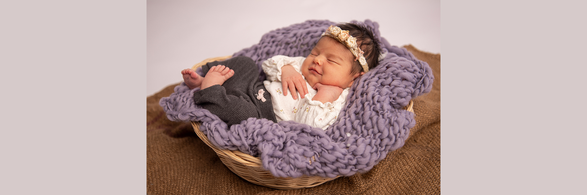 Newbornbaby das laechelt in Korb mit Blumenstrinband auf violetter Kuscheldecke